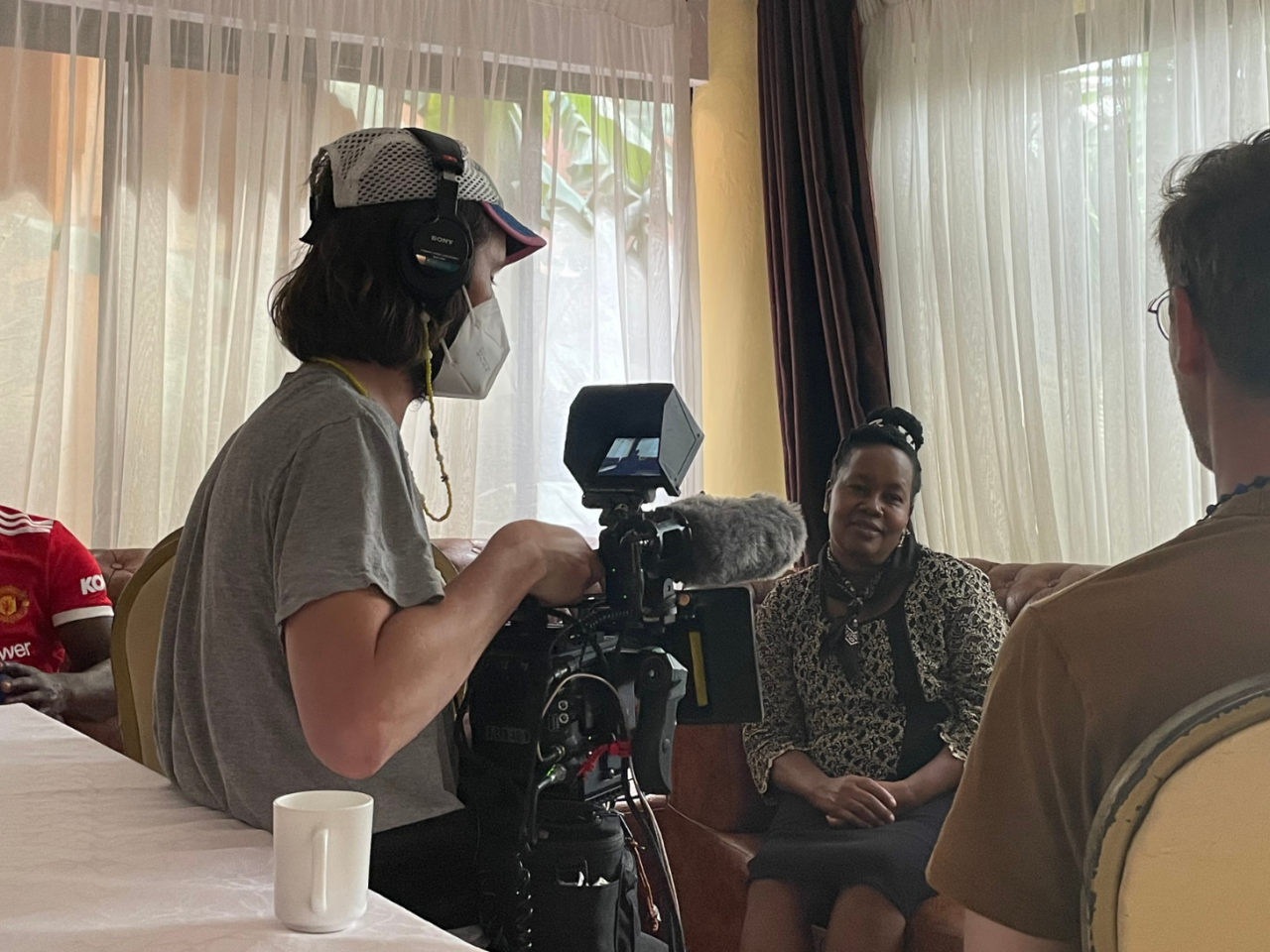 Production still for the film “Rollout.”  Filmmaker Trevor Zimmer and his cinematographer, who are both white, film an interview with a Kenyan woman.  The cinematographer wears a face mask over his nose and mouth.  On the left, a Kenyan man looks at his phone.  He sits outside of the cinematographer’s shot and is not part of the filmed interview.